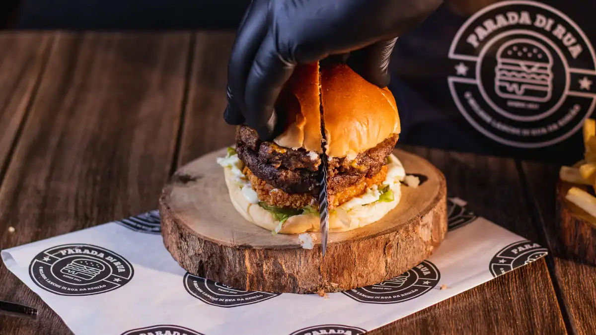 Hambúrguer gourmet com dupla camada de carne, frango empanado e queijo derretido, sendo cortado ao meio para revelar suas camadas suculentas. Perfeito para compartilhar ou saborear sozinho